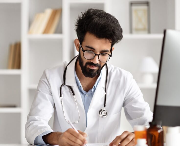 Young bearded indian man wearing medical uniform doctor sitting at desk with computer, taking notes anamnesis, physician consulting woman patient at modern clinic